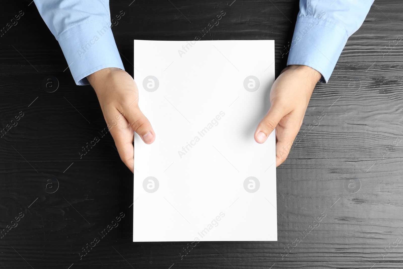 Photo of Man holding blank paper sheet for brochure at black wooden table, top view. Mock up