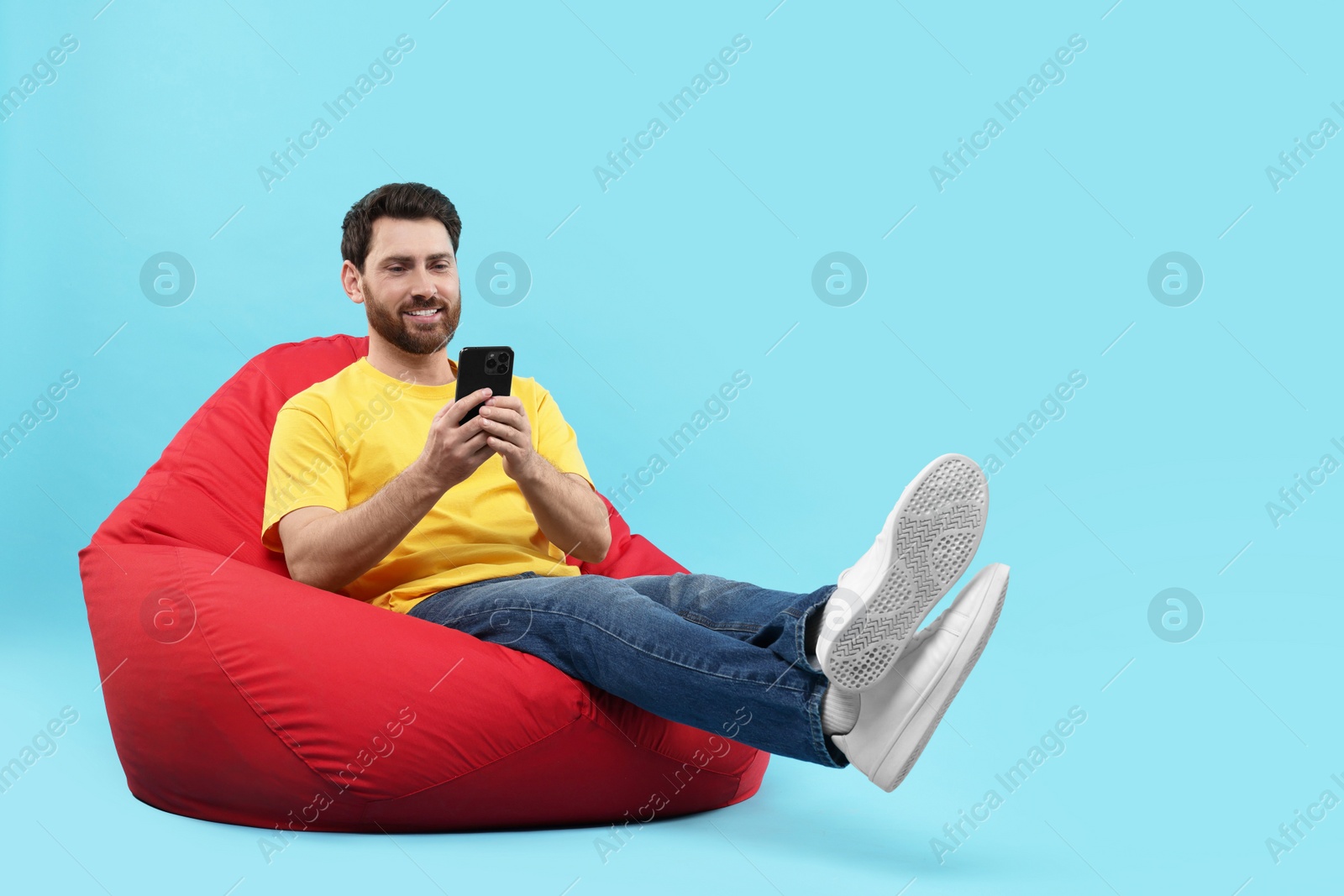 Photo of Smiling man with smartphone sitting on bean bag against light blue background. Space for text