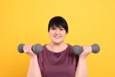 Photo of Happy overweight mature woman doing exercise with dumbbells on yellow background