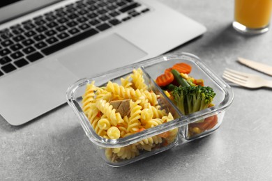 Container with tasty food, laptop and cutlery on light grey table. Business lunch