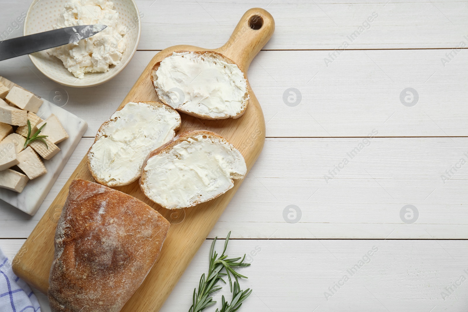 Photo of Slices of baguette with tofu cream cheese and rosemary on white wooden table, flat lay. Space for text