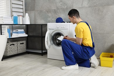 Photo of Young plumber writing results of examining washing machine in bathroom