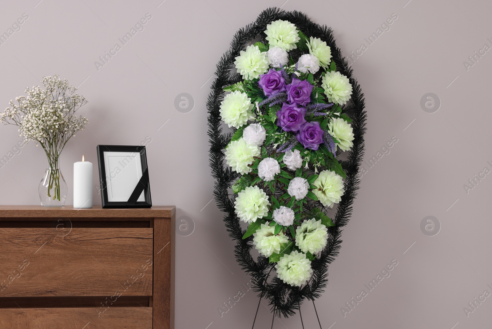 Photo of Wreath of plastic flowers and frame with black ribbon, burning candle on commode in room. Funeral attributes