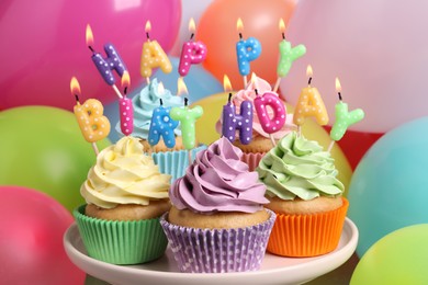 Photo of Birthday cupcakes with burning candles on stand near color balloons, closeup