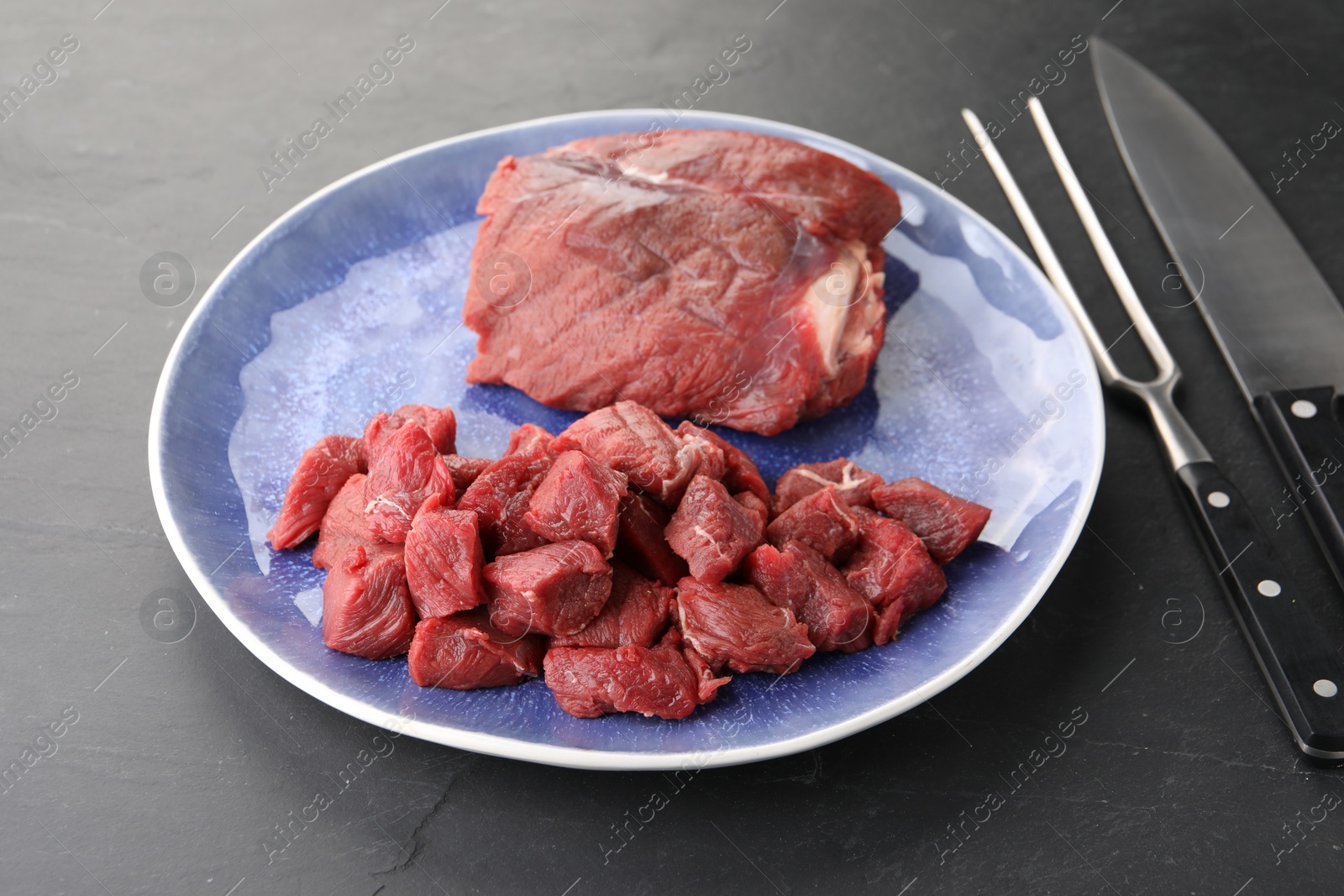 Photo of Plate with pieces of raw beef meat, knife and fork on grey textured table, closeup