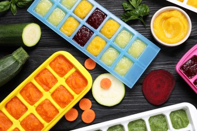 Photo of Different frozen purees in ice cube trays and ingredients of black wooden table, flat lay. Ready for freezing
