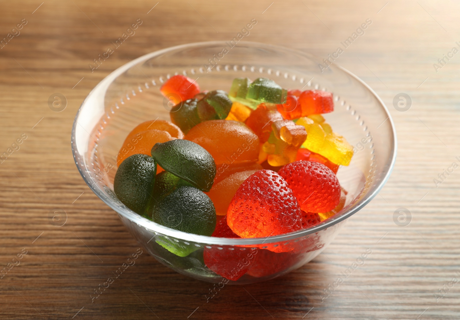 Photo of Different delicious gummy candies in glass bowl on wooden table