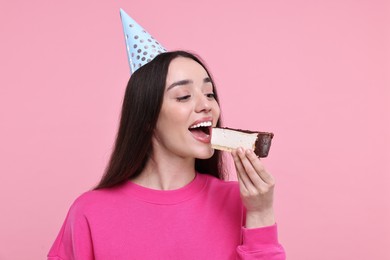 Photo of Woman in party hat eating piece of tasty cake on pink background
