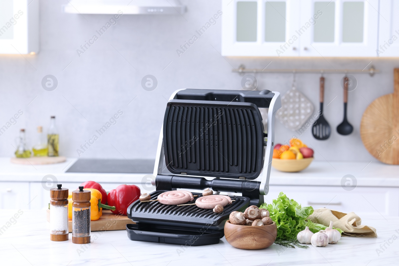 Photo of Electric grill with homemade sausages, mushrooms, vegetables and spices on white table in kitchen