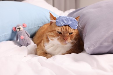 Photo of Cute ginger cat with sleep mask and crocheted mouse resting on bed