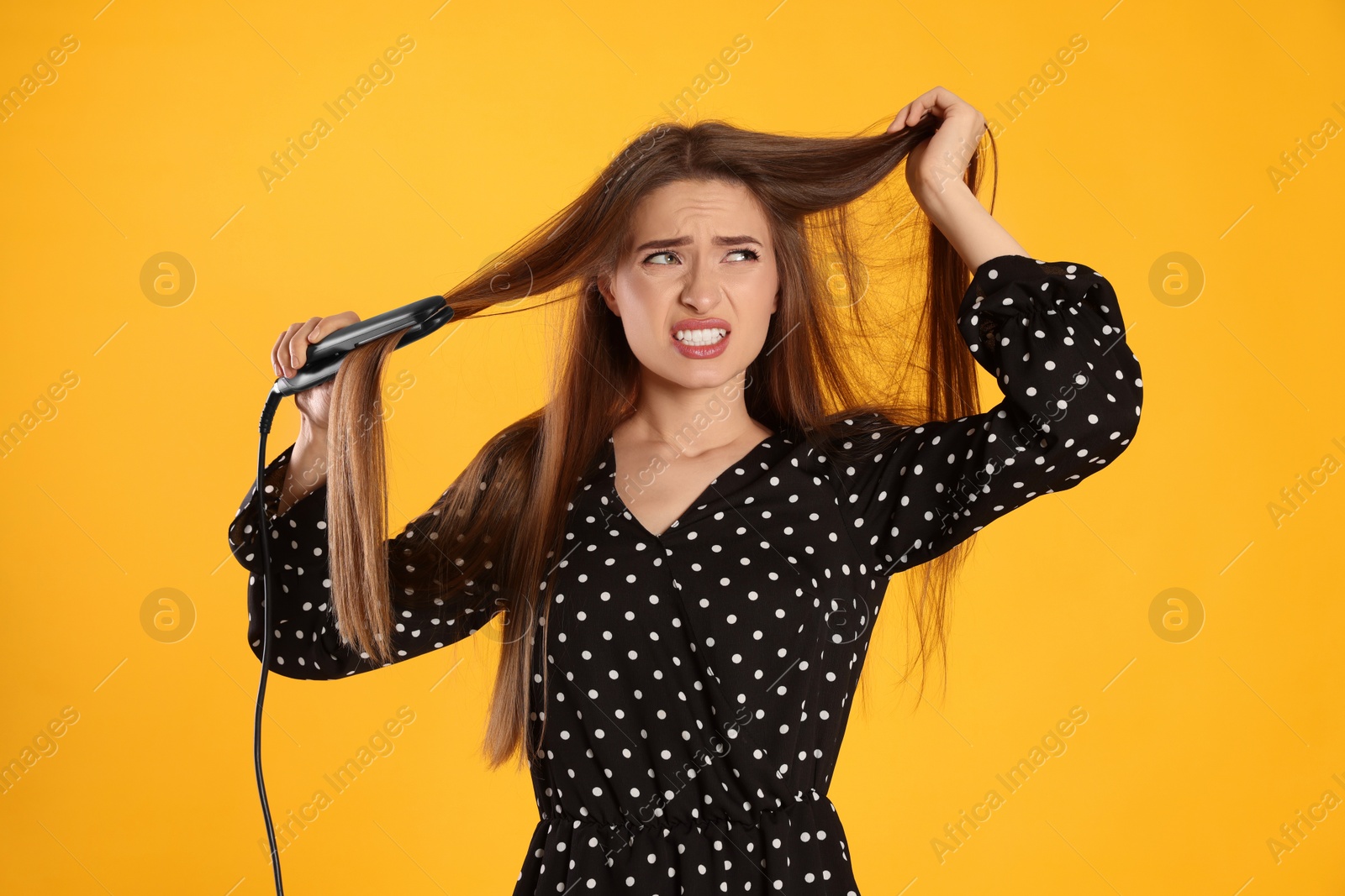 Photo of Stressed young woman with flattening iron on yellow background. Hair damage