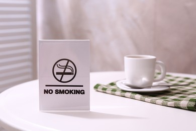 Photo of No Smoking sign and cup of coffee on white table indoors