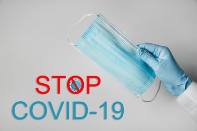 Doctor holding medical mask near text Stop Covid-19 on light background, closeup. Protective measures during pandemic