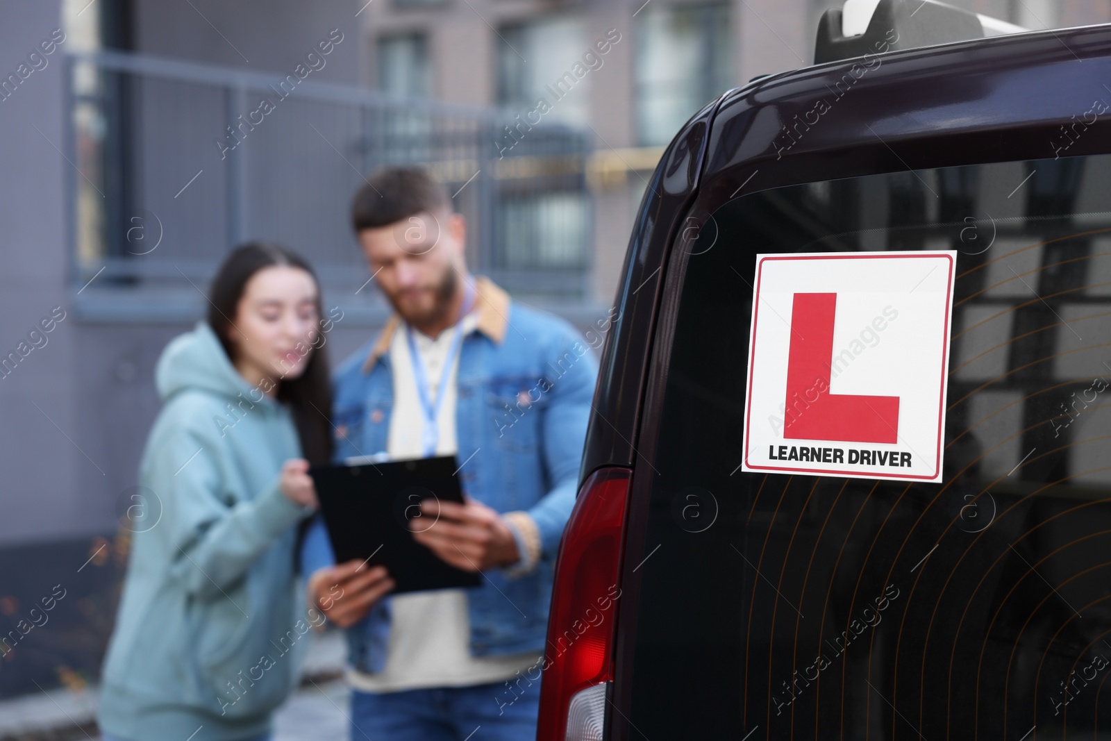 Photo of Learner driver and instructor with clipboard near car outdoors, selective focus on L-plate. Driving school