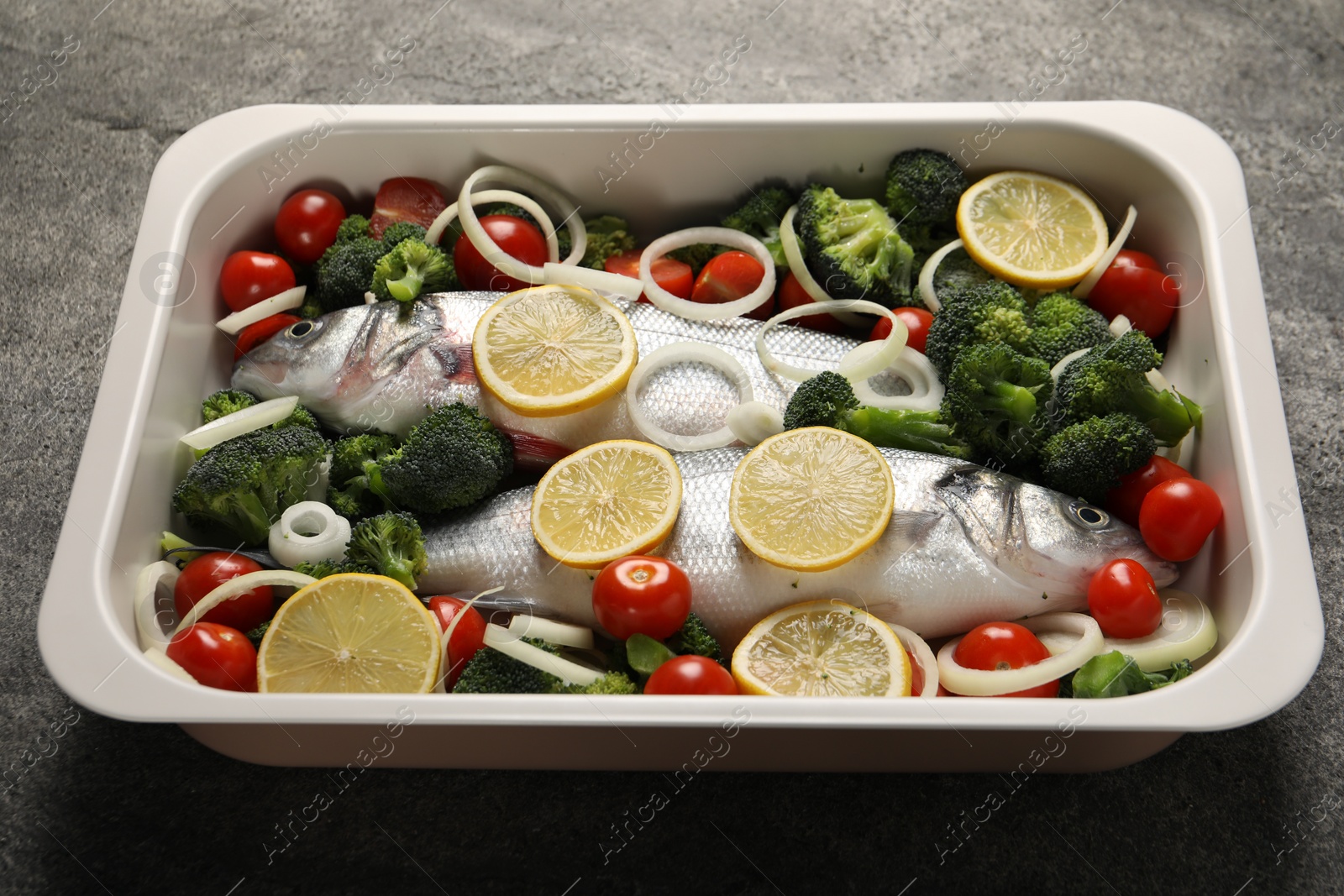 Photo of Raw fish with vegetables and lemon in baking dish on grey textured table