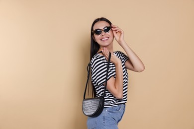 Beautiful young woman with stylish bag in sunglasses on beige background