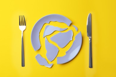 Pieces of broken ceramic plate and cutlery on yellow background, flat lay