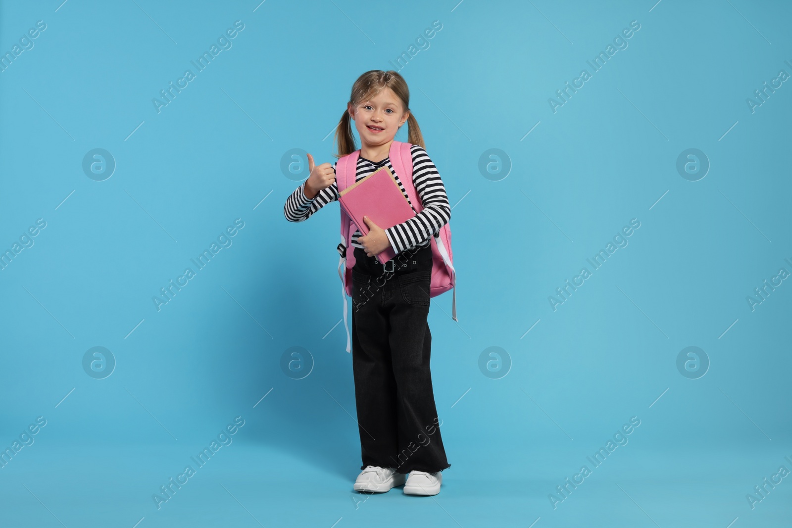 Photo of Happy schoolgirl with backpack and books showing thumb up gesture on light blue background