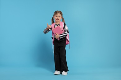 Happy schoolgirl with backpack and books showing thumb up gesture on light blue background