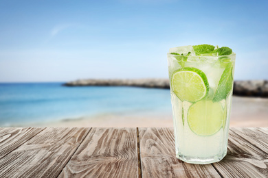 Image of Tasty lemonade with ice cubes and lime on wooden table near sea, space for text