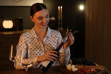 Romantic dinner. Happy woman opening wine bottle with corkscrew at table indoors