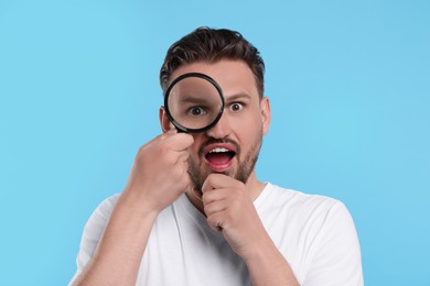 Emotional man looking through magnifier on light blue background