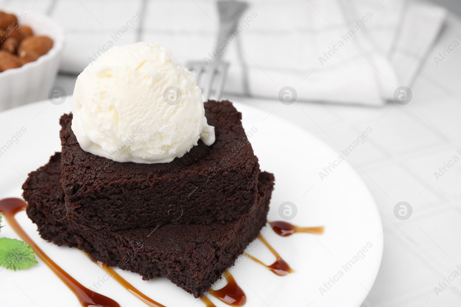 Photo of Tasty brownies served with ice cream and caramel sauce on white tiled table, closeup