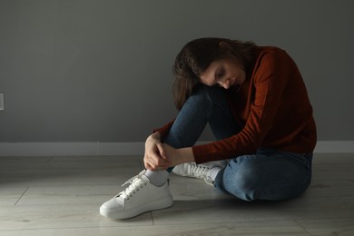 Sad young woman sitting on floor near grey wall indoors