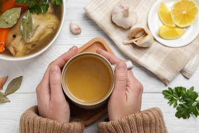 Woman with cup of hot delicious bouillon at white wooden table, top view