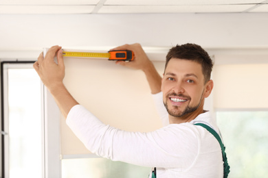 Handyman with tape measure installing roller window blind indoors