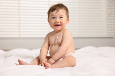 Photo of Cute little baby with moisturizing cream onto body sitting on bed at home