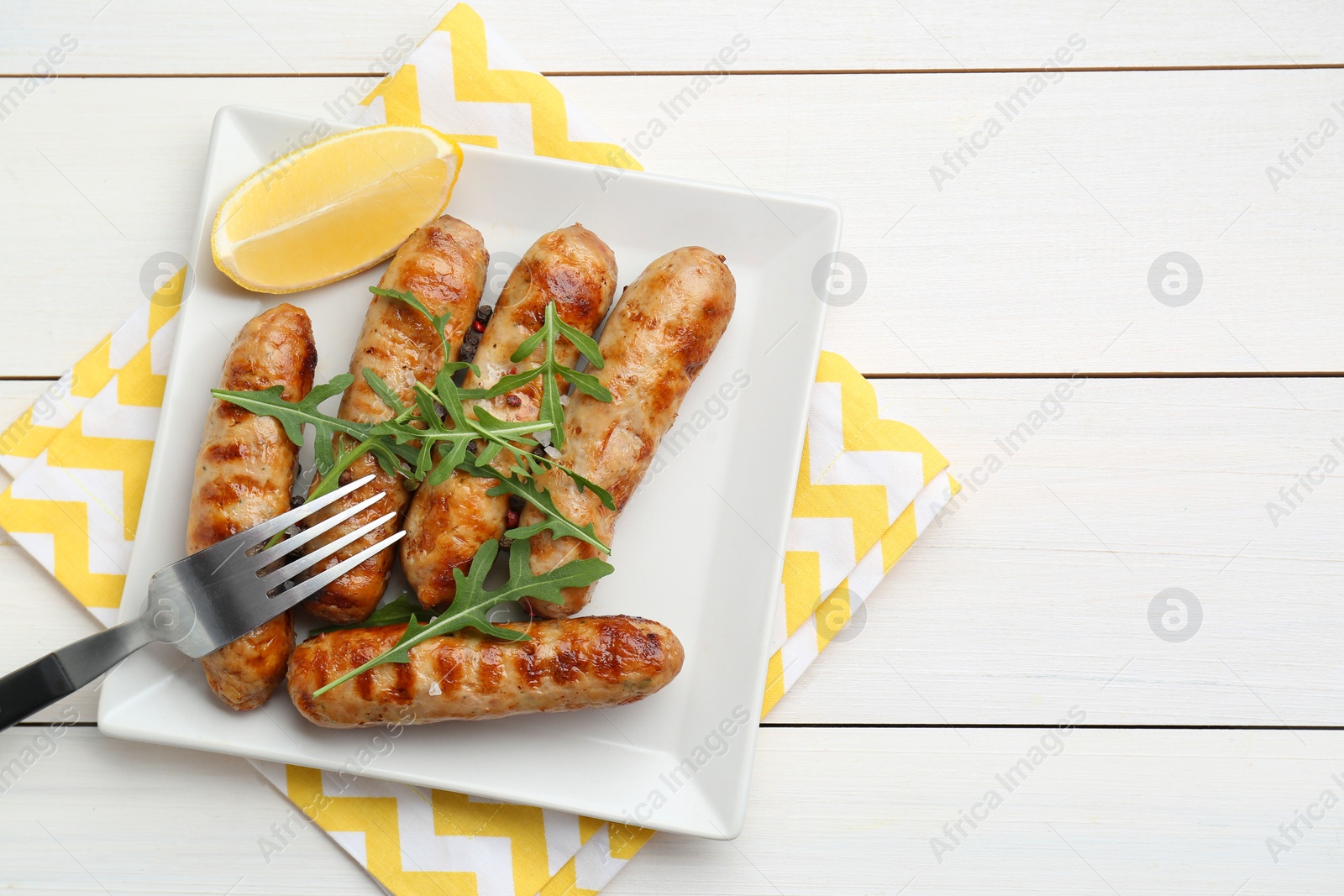 Photo of Tasty grilled sausages served with lemon and arugula on white wooden table, top view. Space for text