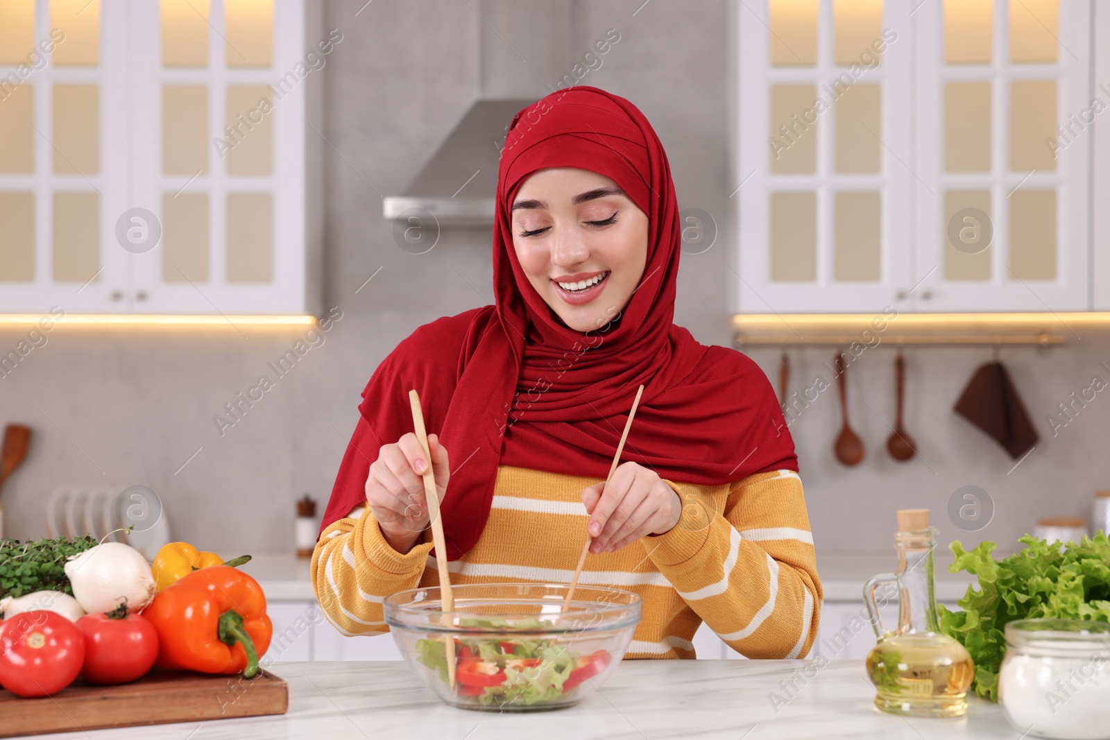 Photo of Muslim woman making delicious salad with vegetables at white table in kitchen