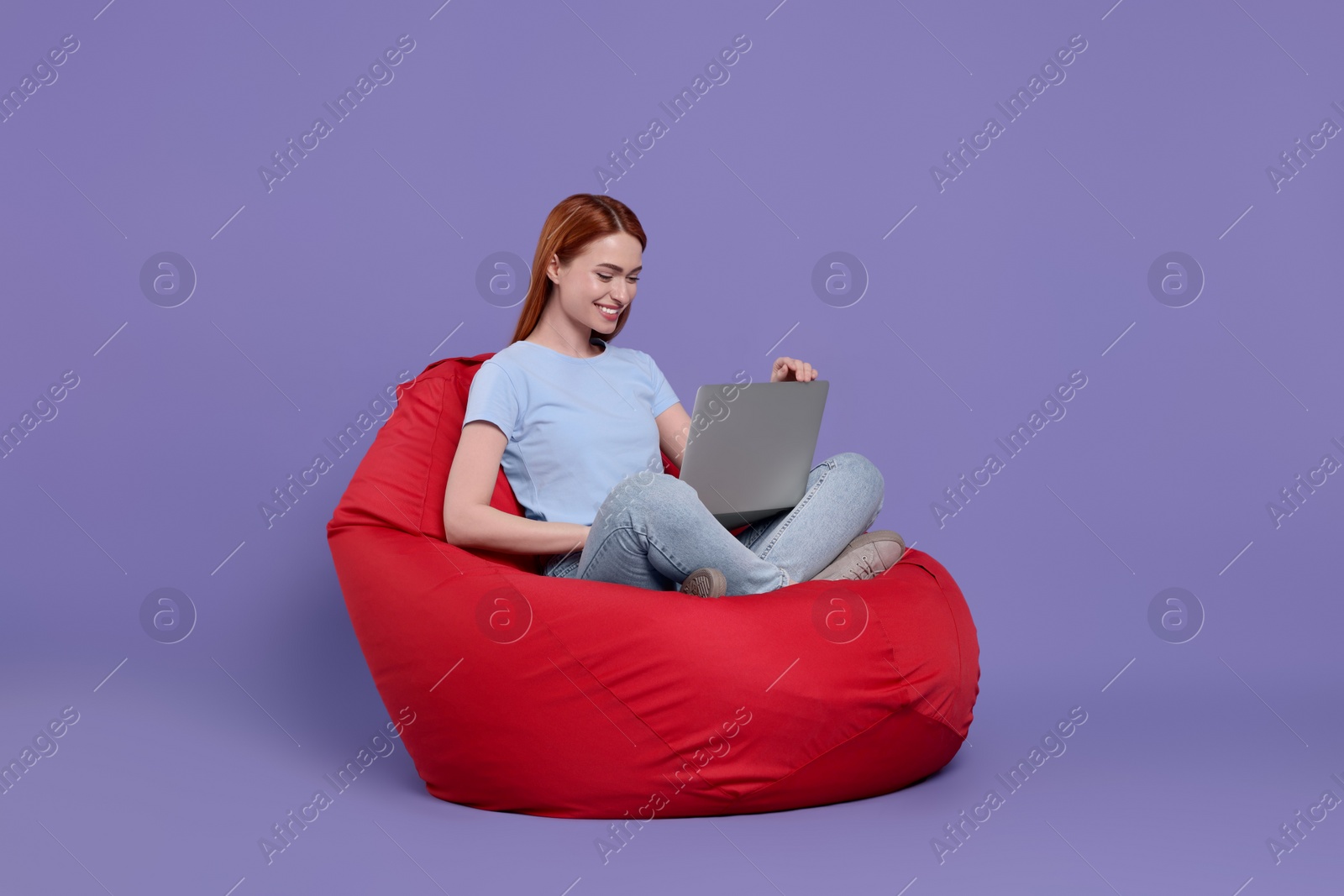 Photo of Smiling young woman with laptop sitting on beanbag chair against lilac background, space for text