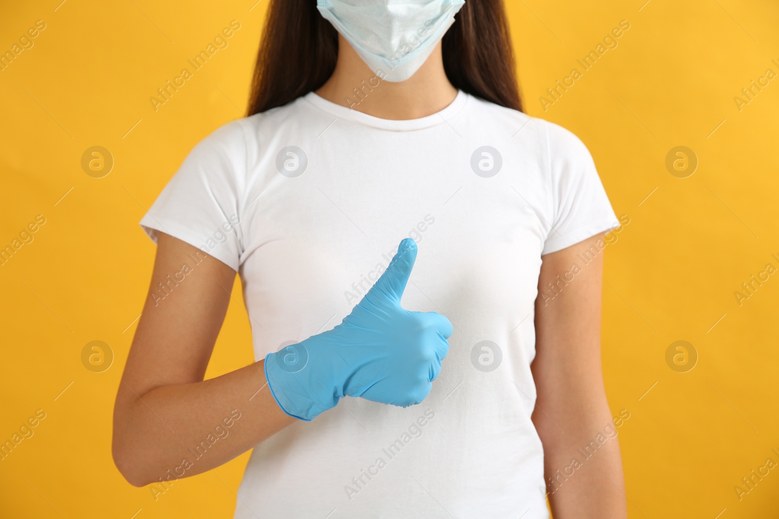 Photo of Woman in protective face mask and medical gloves showing thumb up gesture on yellow background, closeup