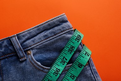 Jeans and measuring tape on orange background, top view. Weight loss concept