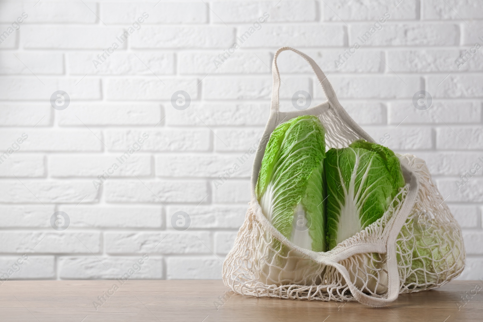 Photo of Fresh Chinese cabbages in string bag on wooden table near white brick wall. Space for text