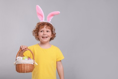 Happy boy in cute bunny ears headband holding basket with Easter eggs on light grey background. Space for text