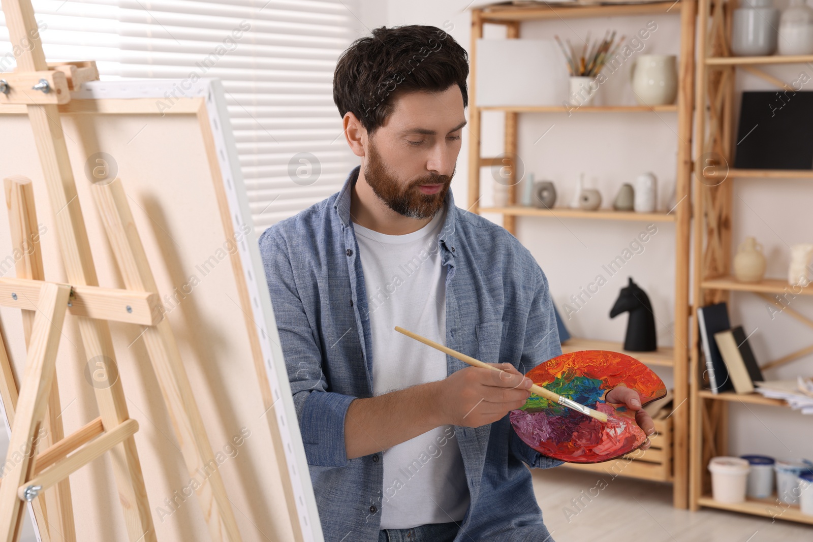 Photo of Man with brush and palette painting in studio. Using easel to hold canvas
