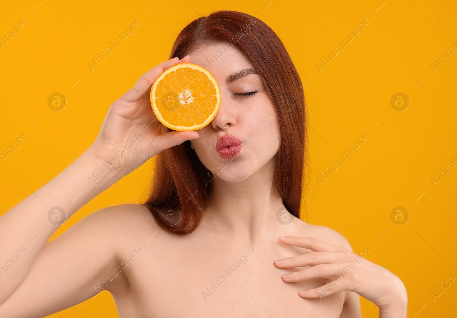 Photo of Beautiful woman with freckles covering eye with half of orange and sending air kiss on yellow background