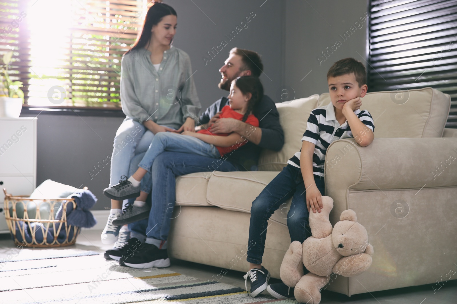 Photo of Unhappy little boy feeling jealous while parents spending time with his sister at home