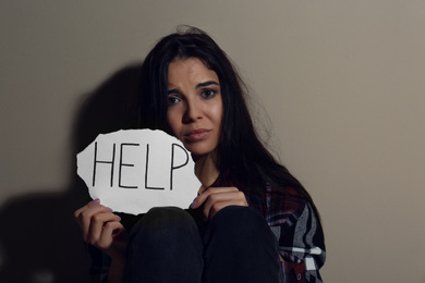 Abused young woman with sign HELP near beige wall. Domestic violence concept