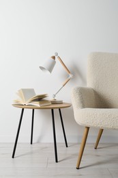 Photo of Stylish lamp with open book on wooden coffee table and soft armchair in light room. Interior design