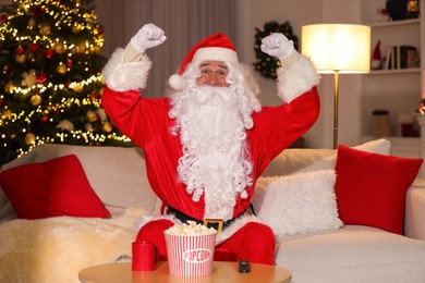 Photo of Merry Christmas. Emotional Santa Claus watching TV on sofa at home