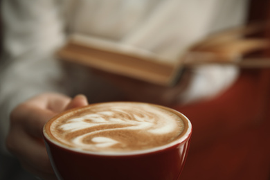 Woman with coffee reading book indoors, focus on cup