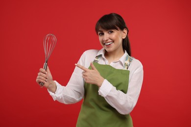 Photo of Happy professional confectioner in apron pointing at whisk on red background
