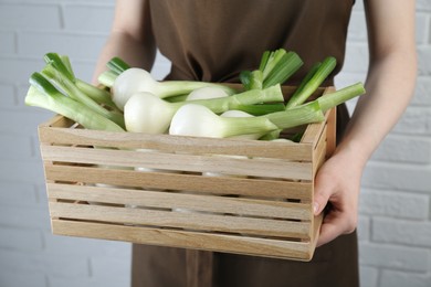 Photo of Woman holding wooden crate with green spring onions near white brick wall, closeup