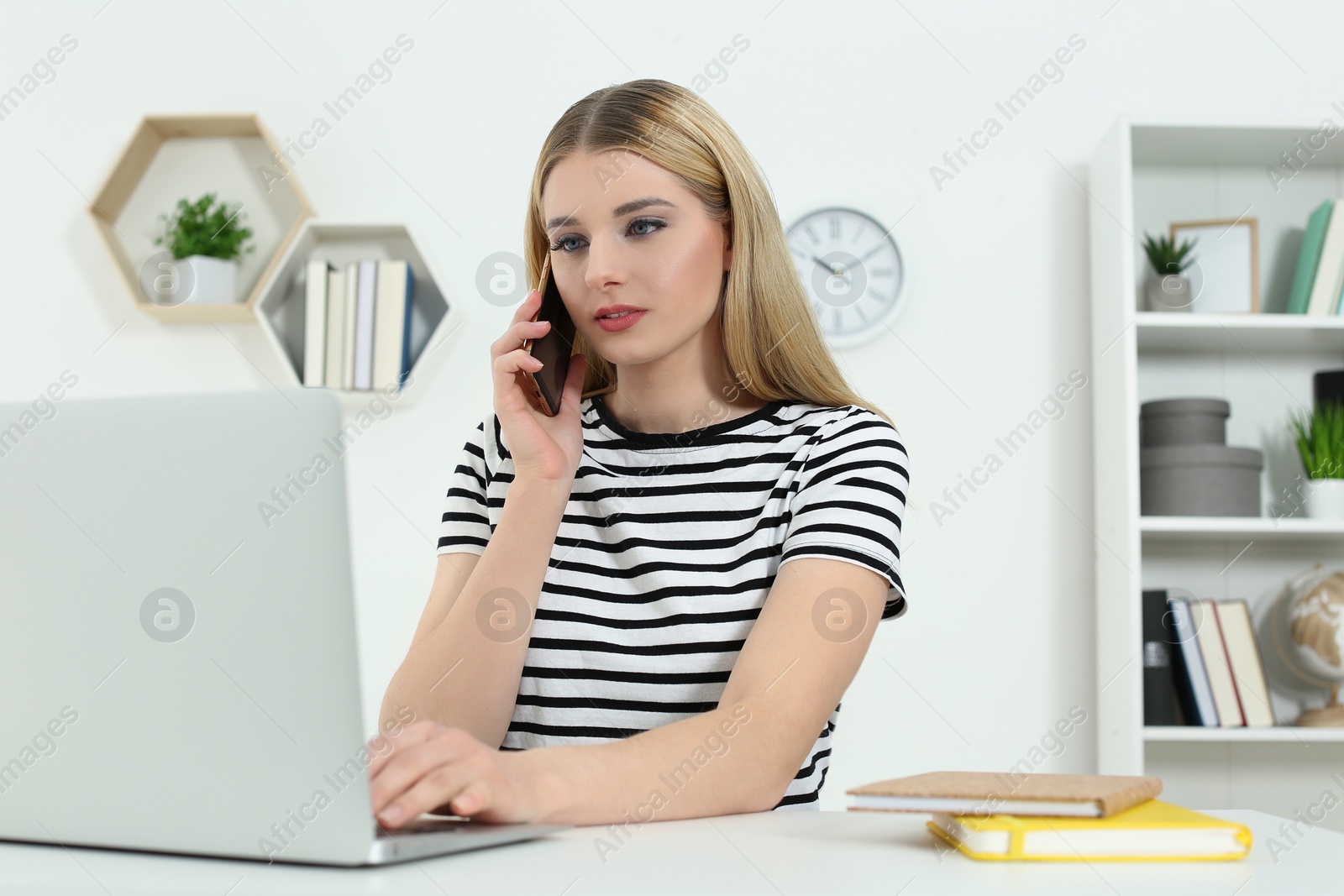 Photo of Home workplace. Woman talking on smartphone near laptop at white desk in room