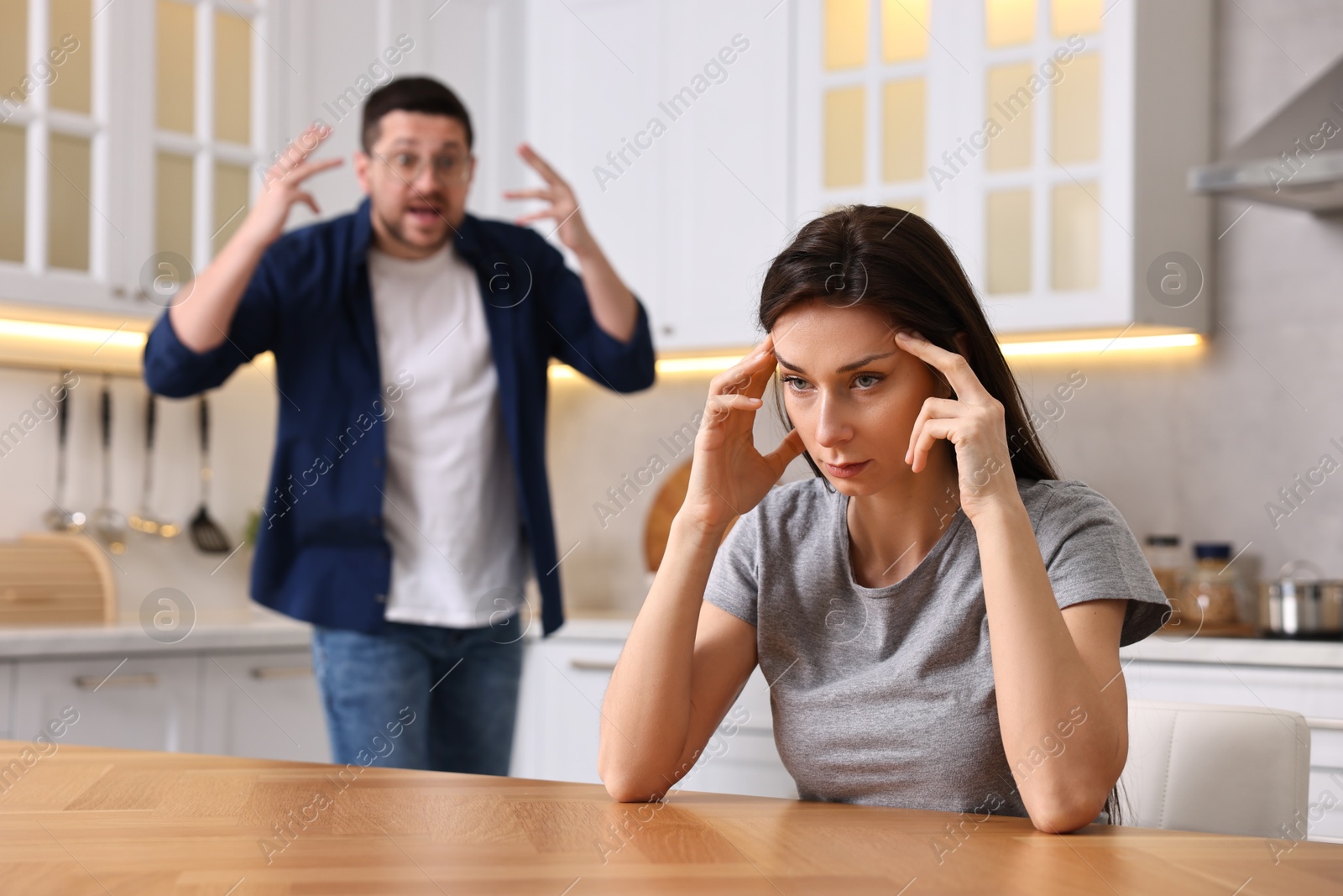 Photo of Stressed wife sitting at table while her angry husband screaming at her in kitchen, selective focus. Relationship problems