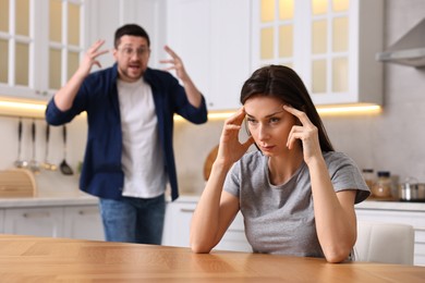 Stressed wife sitting at table while her angry husband screaming at her in kitchen, selective focus. Relationship problems